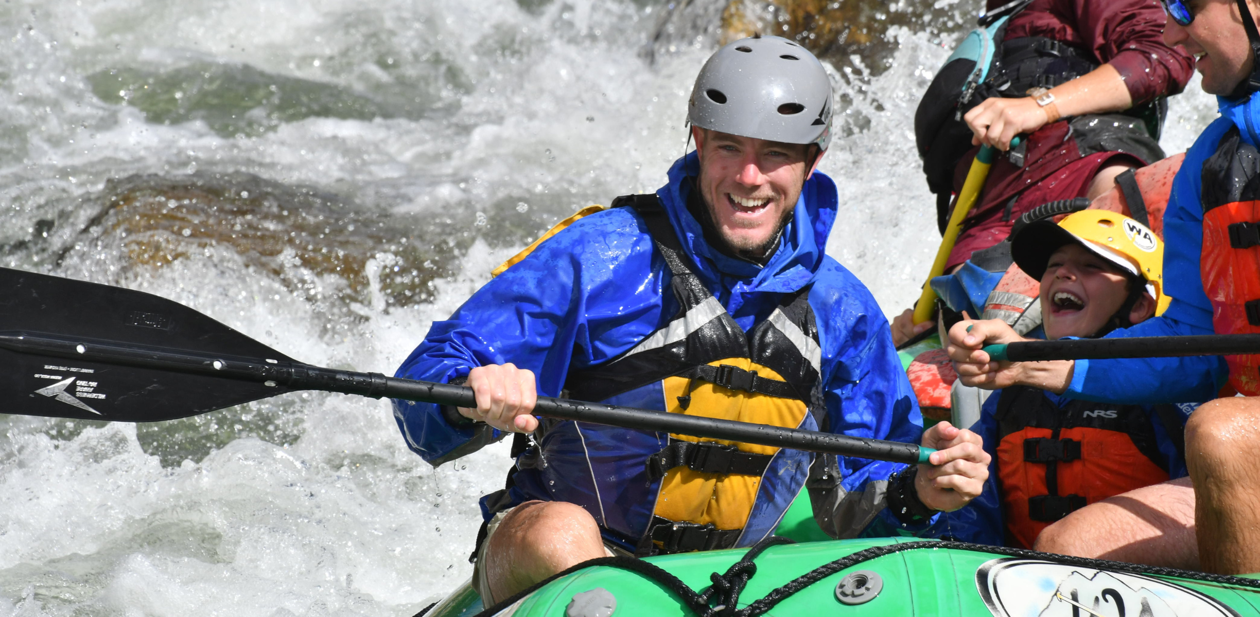 A father and son smile an laugh as they navigate white water in a green raft