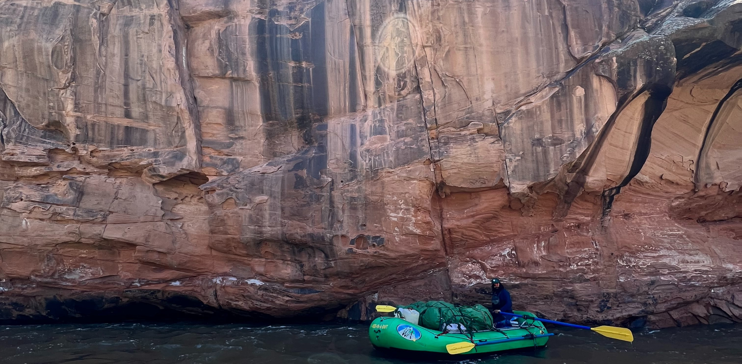 A green raft stops under a large red rock wall