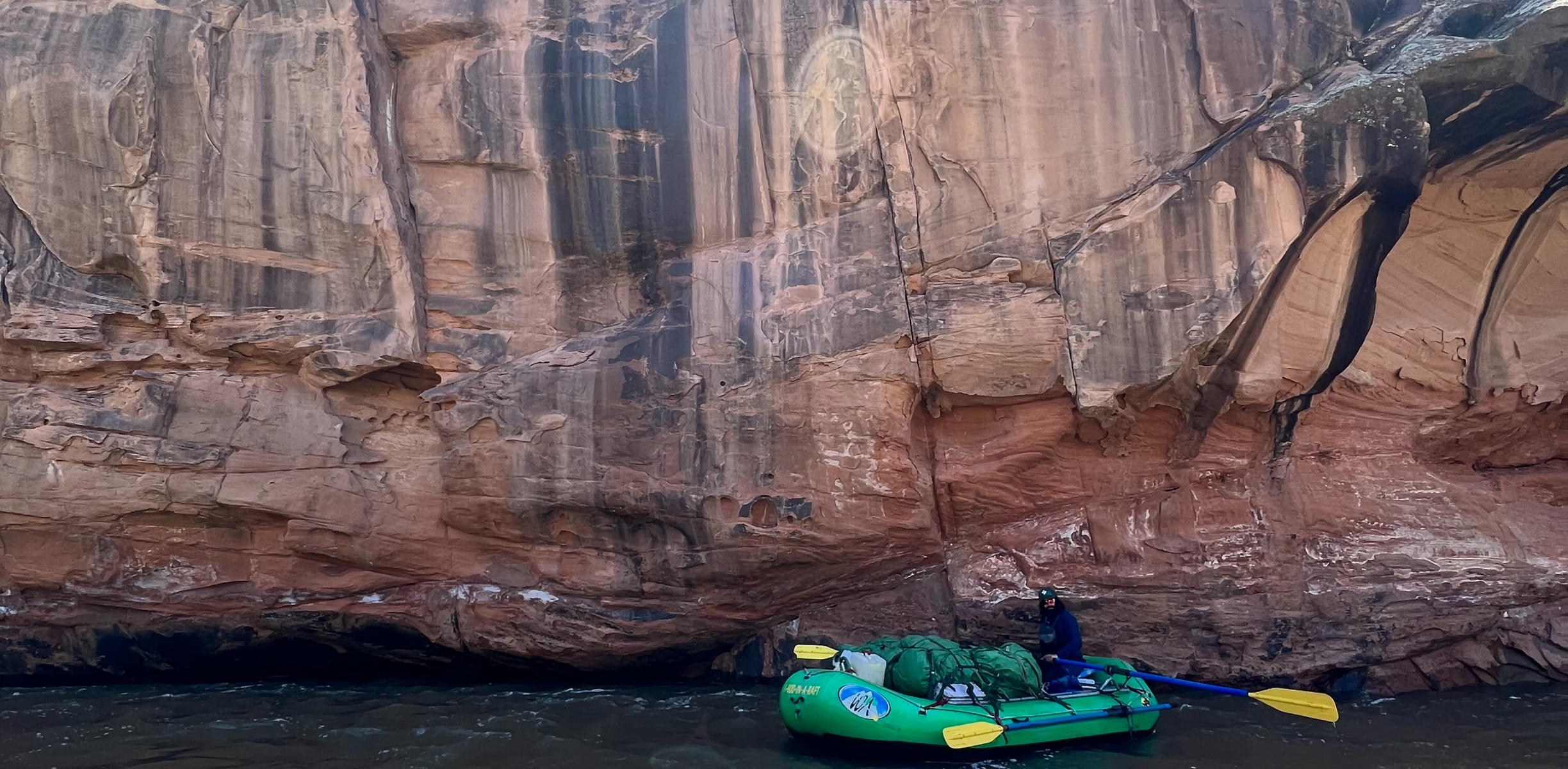 A green raft sits on a river under a red rock wall