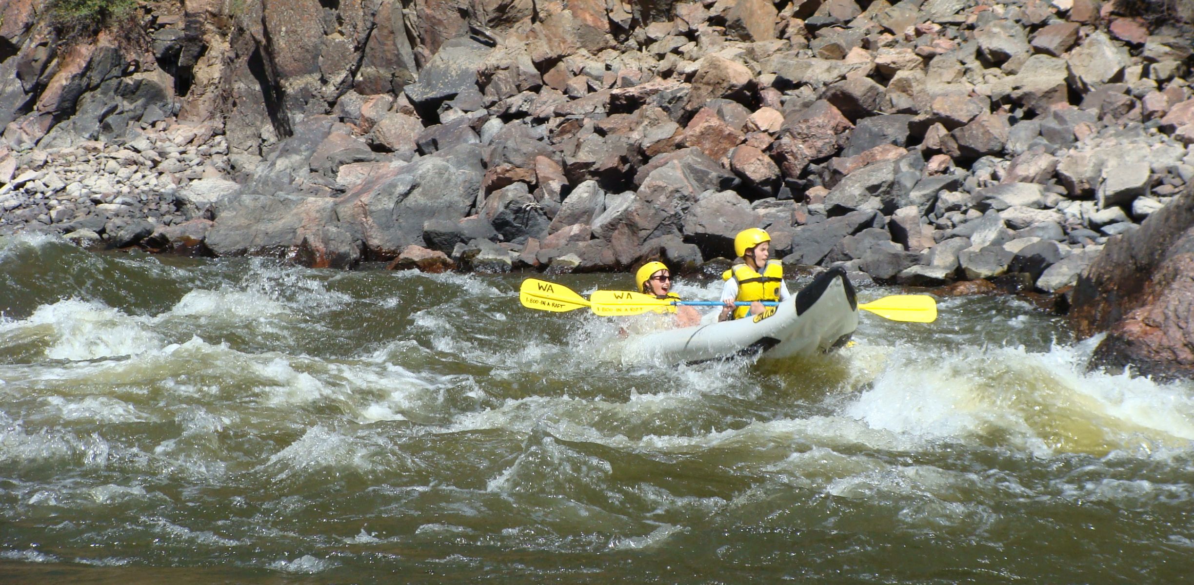 Two kids in a white ducky paddle through river rapids