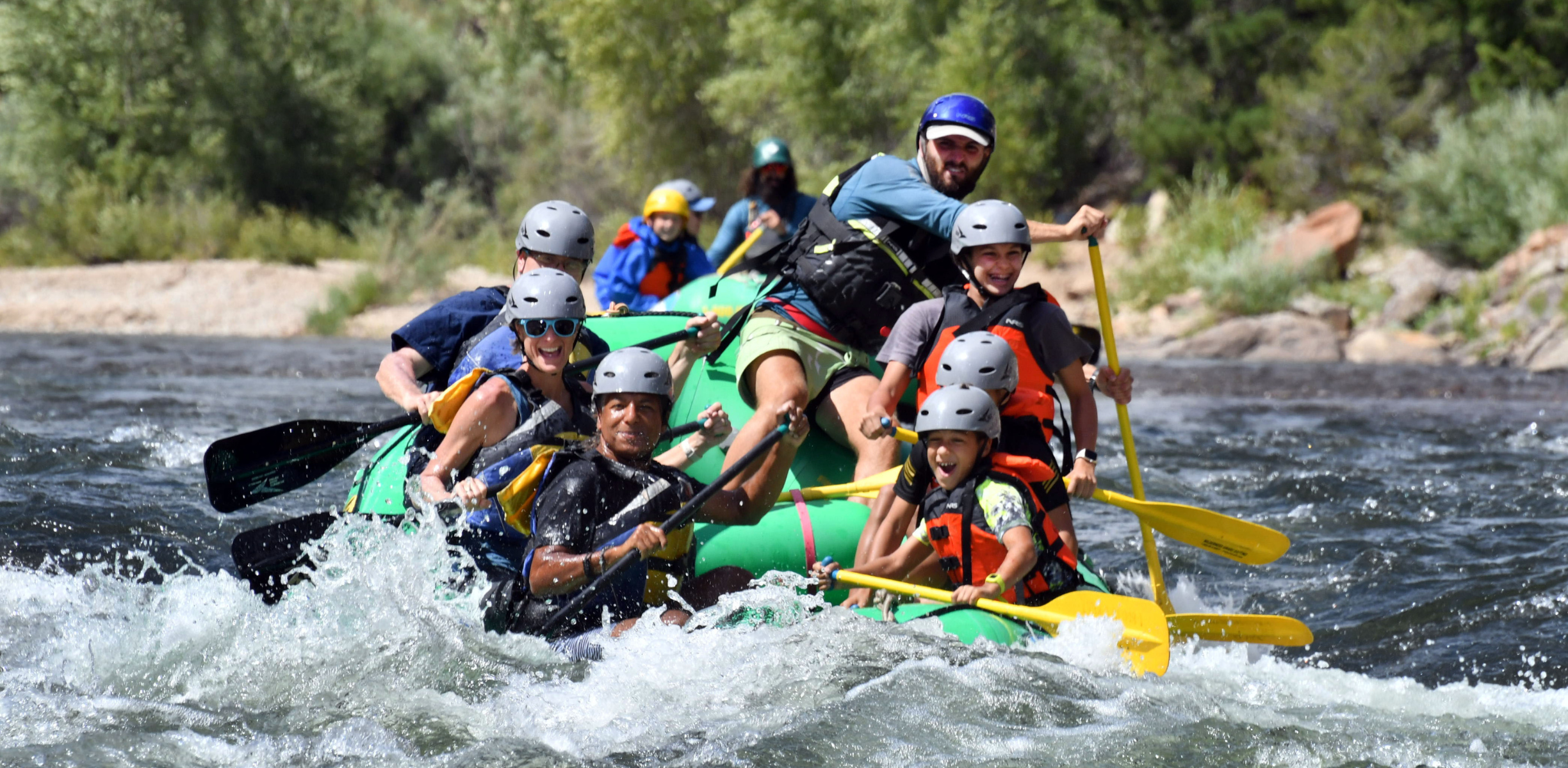 A family on a green raft on the river laugh as they paddle through rapids