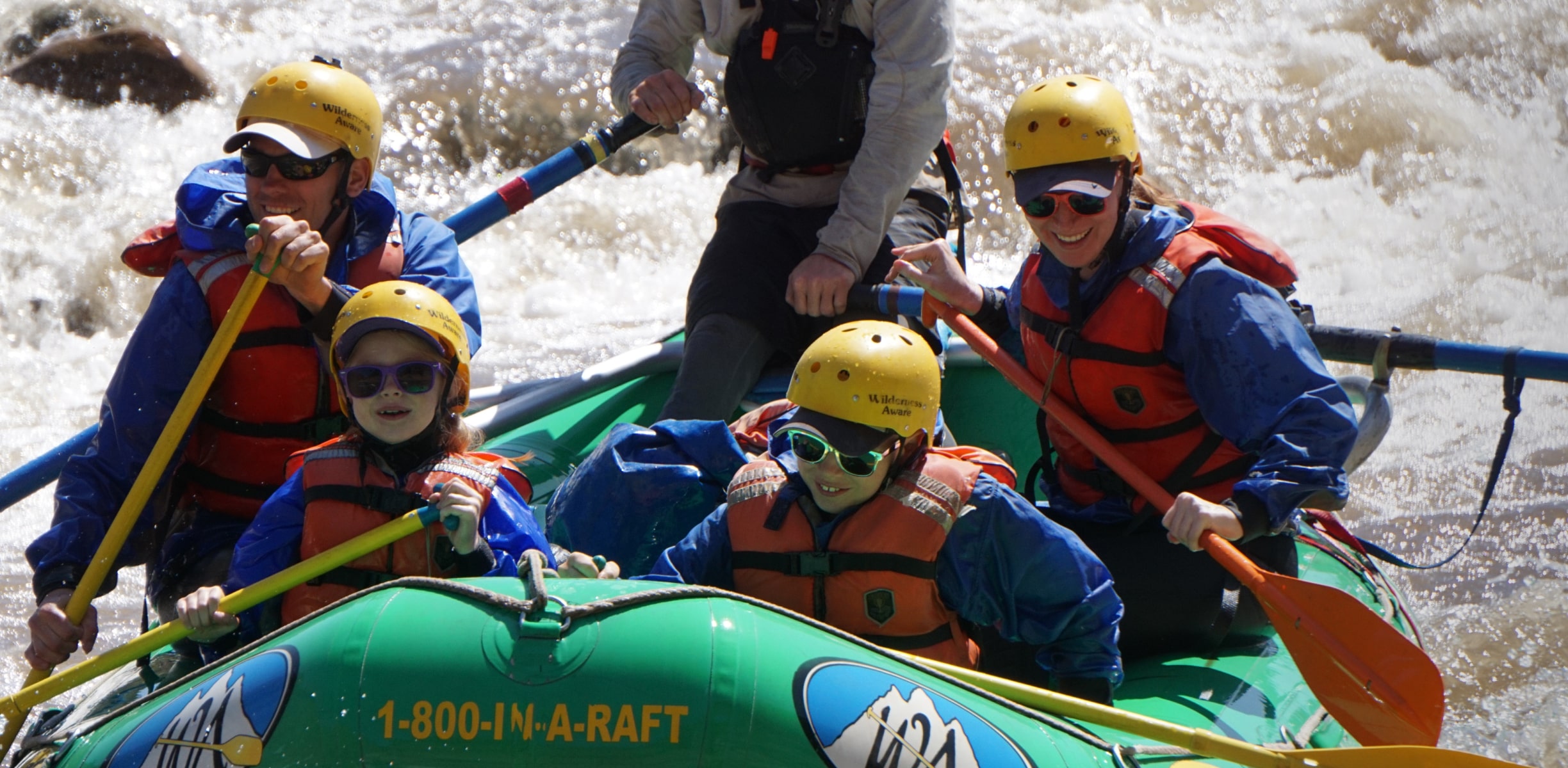 A family white water rafting with orange life vests and yellow helmets