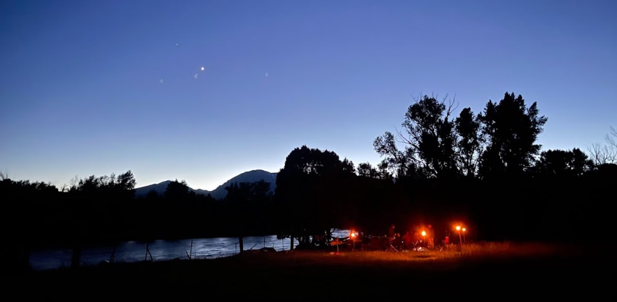 River camp at night with campers gathered around a glowing campfire, illuminated by string lights, during an overnight rafting trip in Colorado.