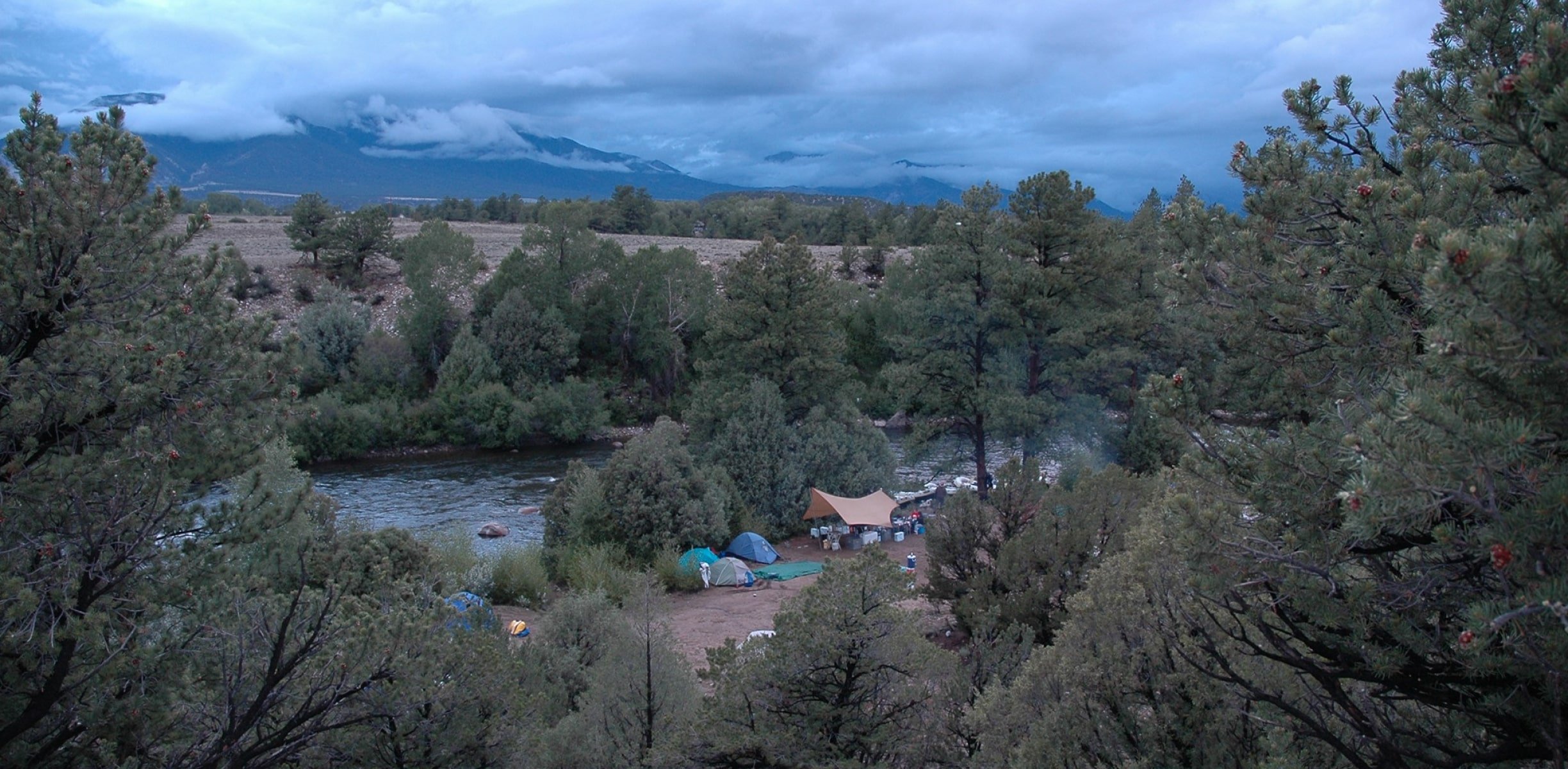 Campers set up tents along the riverbank with a distant mountain under cool blue skies, during an Arkansas River multi-day rafting trip.