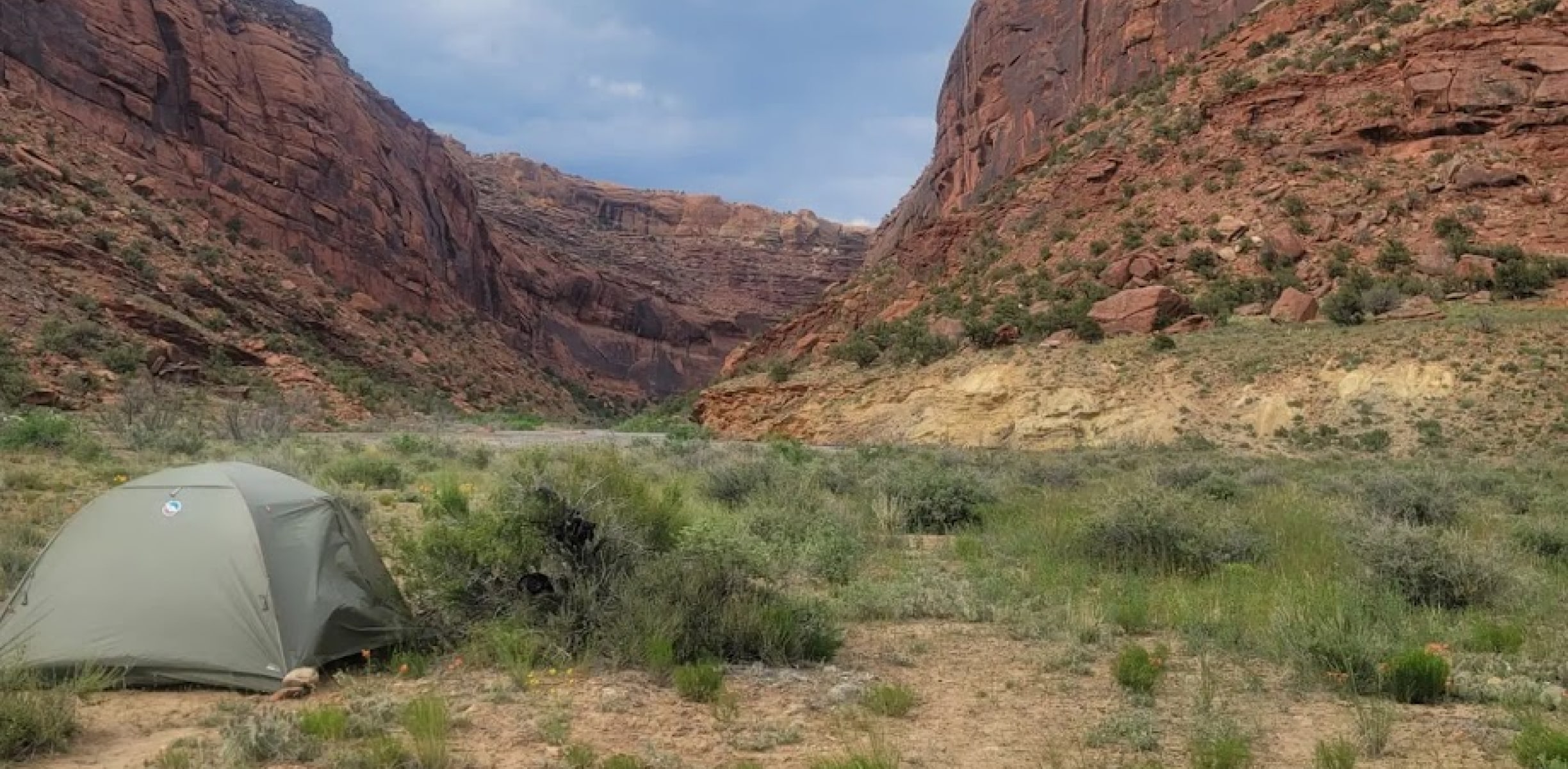 a green tents sits on a river bank next to a river with a scenic red rock canyon in the distance