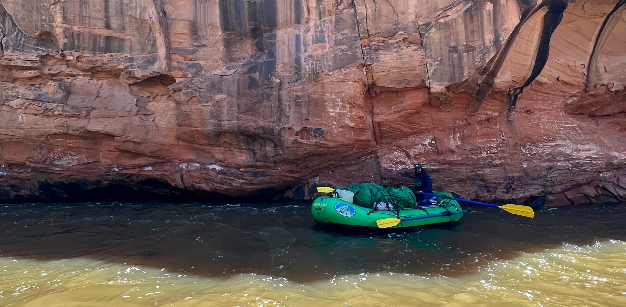 A raft guide sits in his raft next to a large red rock wall on the Dolores River