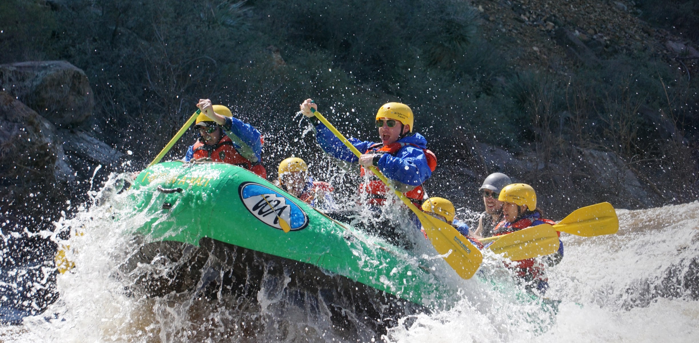 A green raft with a group of rafters with yellow paddles hit a big rapid