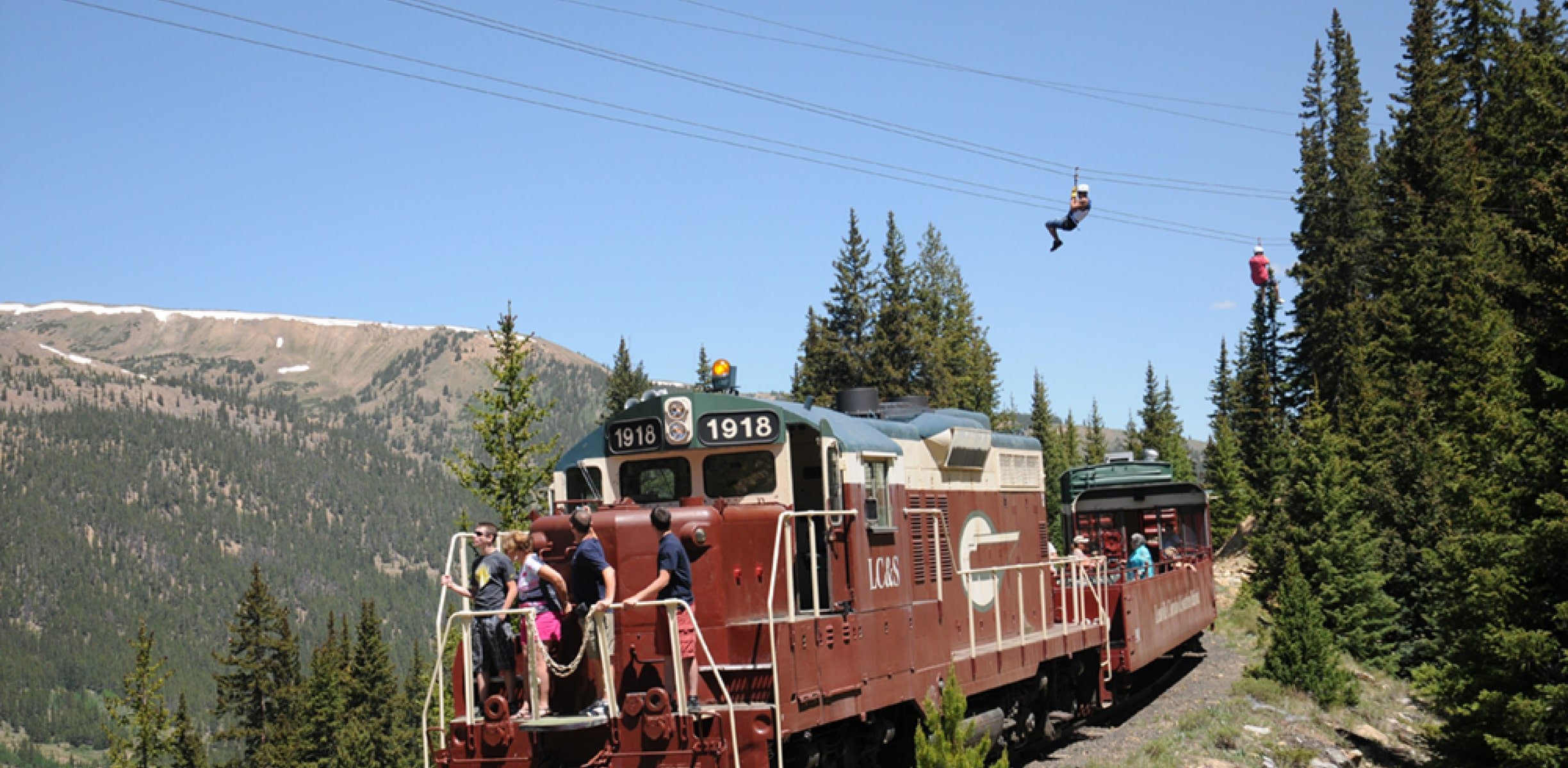 Two people zip lining while a train runs underneath.