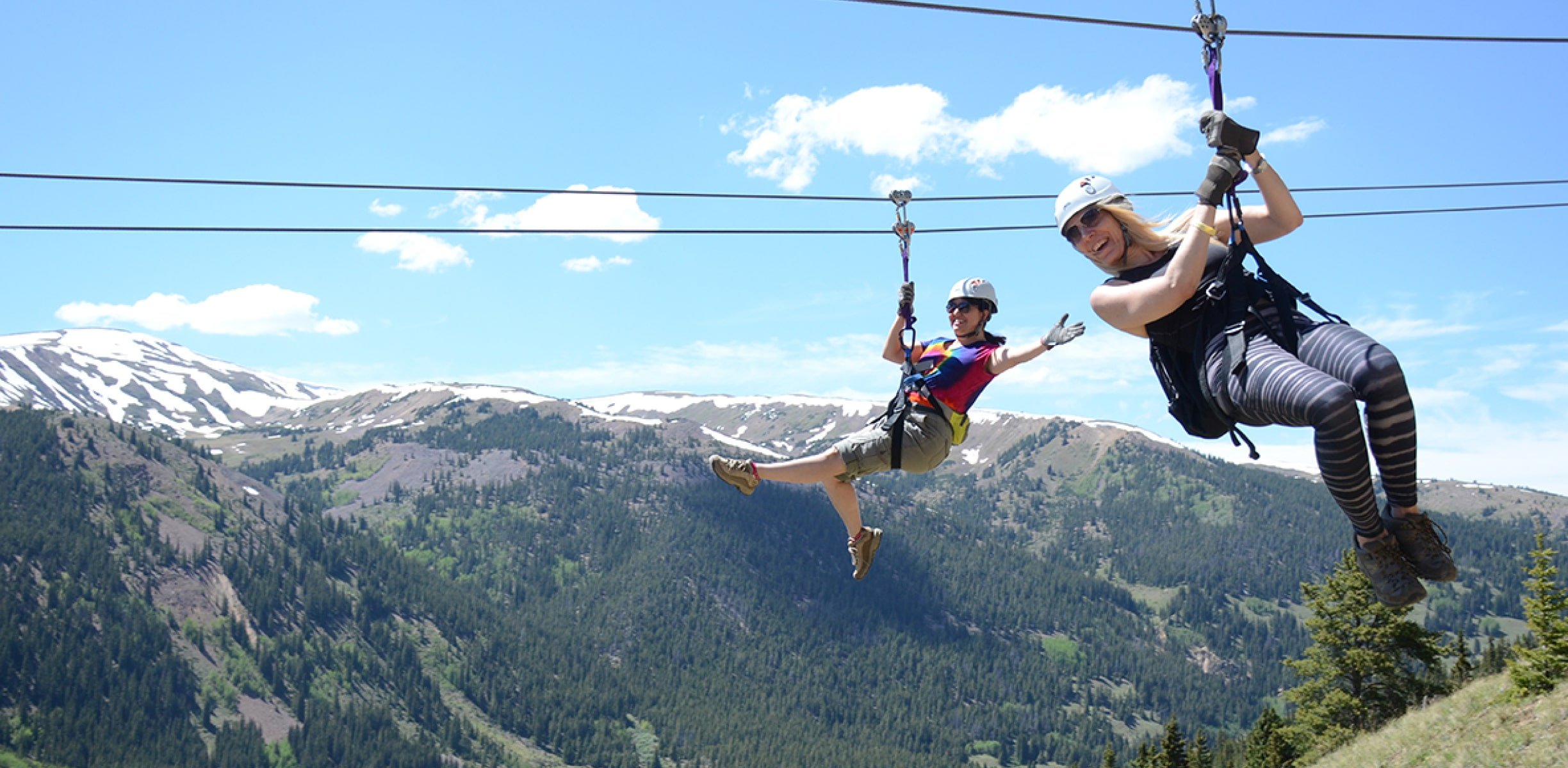 Two happy people ziplining with snowy Colorado mountains behind them