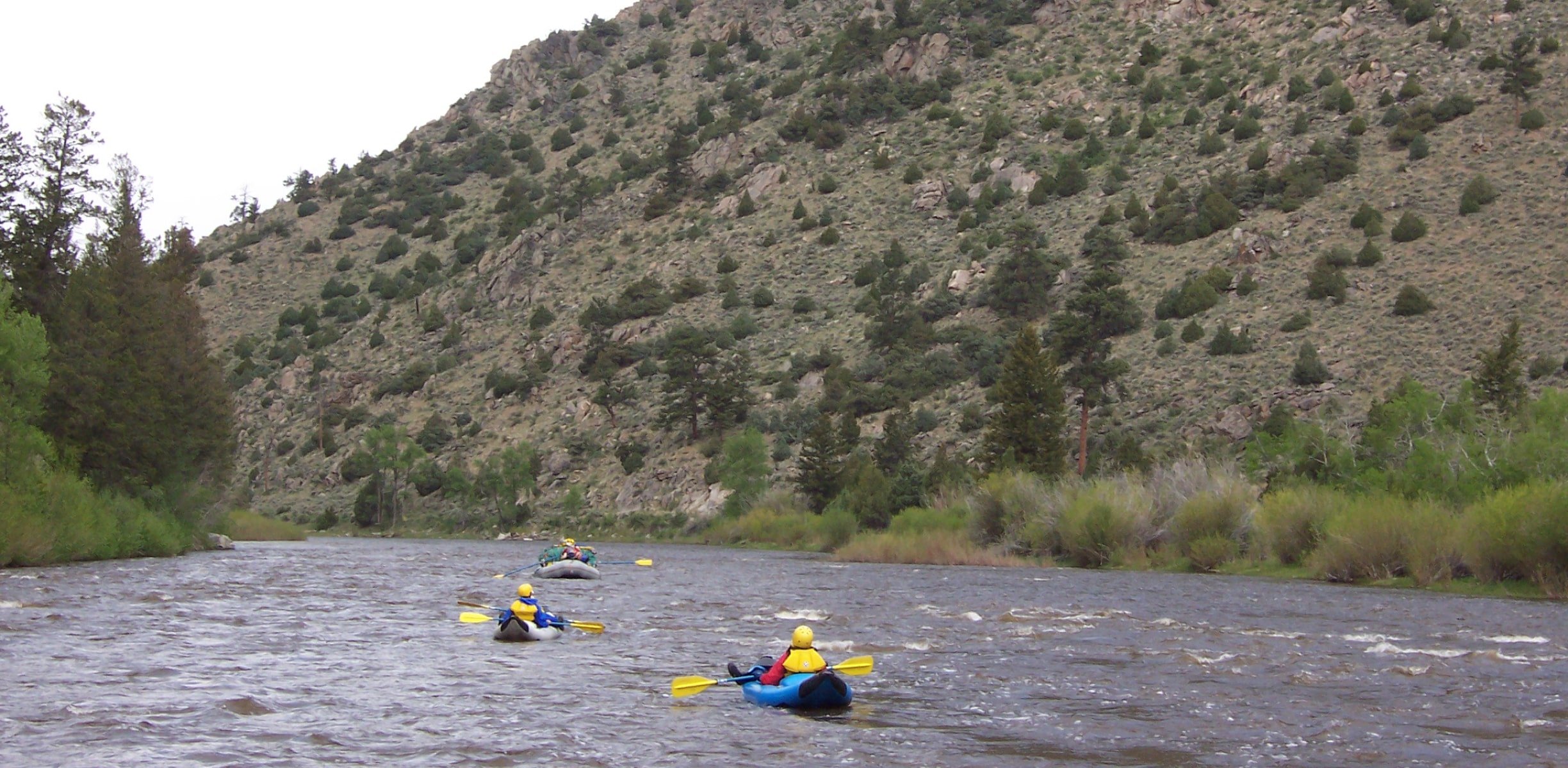 People paddling in inflatable kayaks on a river under a rocky mountain