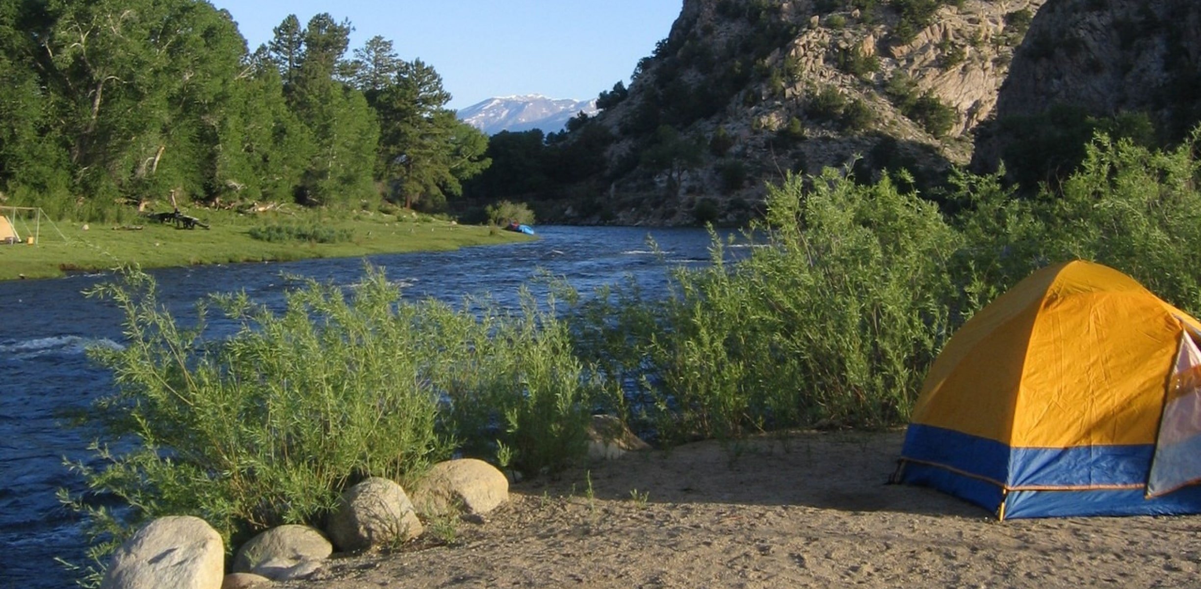 A sunny day with a tent on the river bank in colorado with snowcapped mountains in the background.