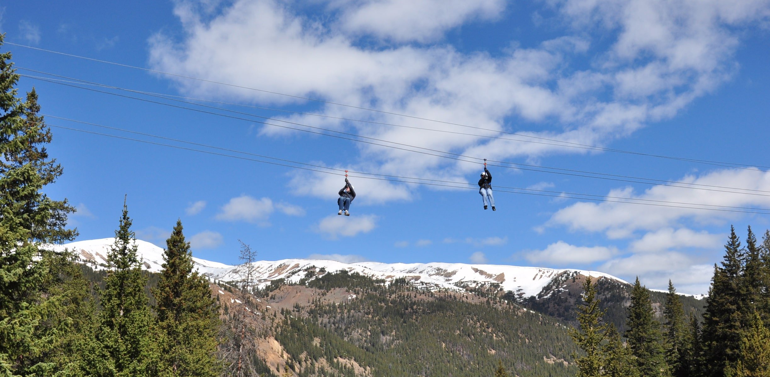 Two people zip lining with snowy Colorado mountains behind them