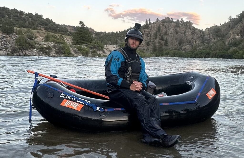 A man sits on a small black raft wearing a dry suit on a river