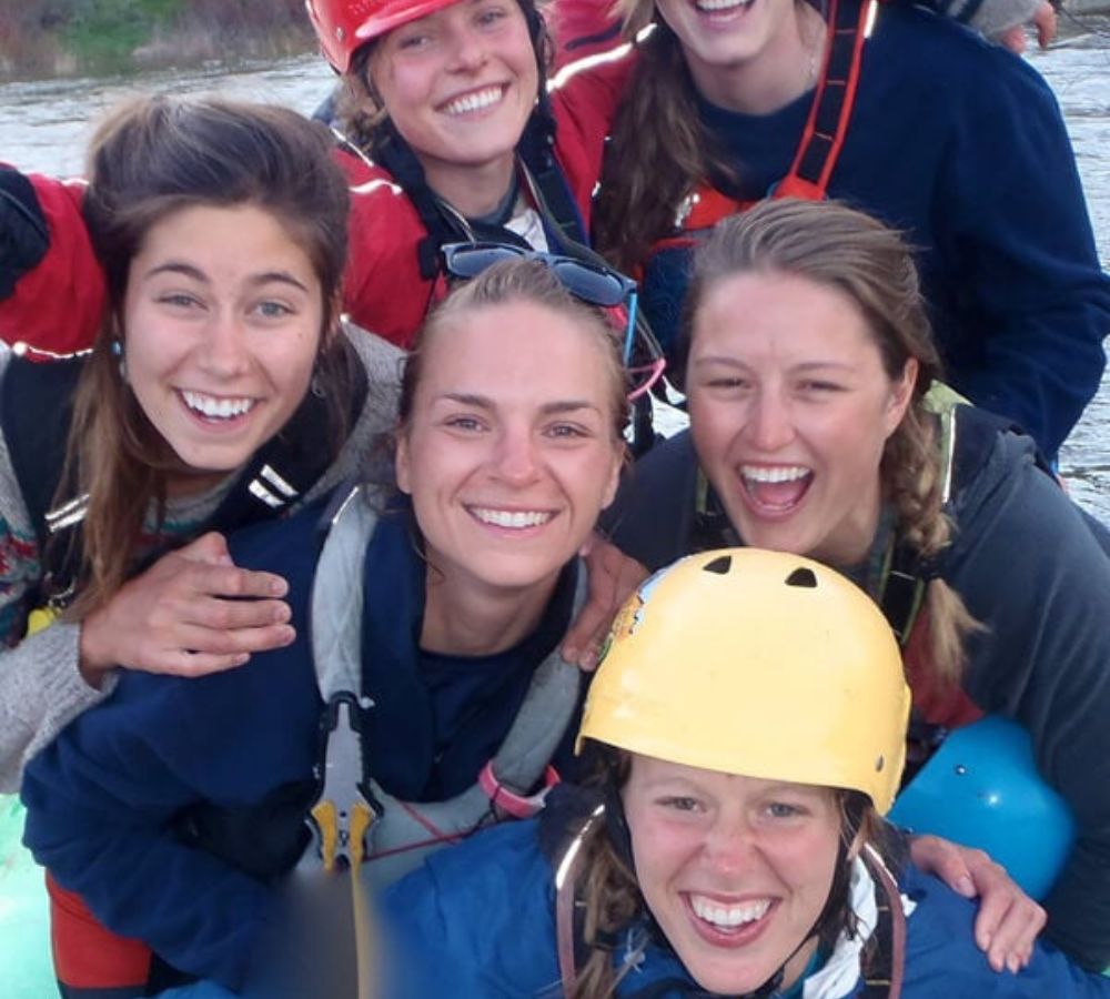 Six women smile and laugh in rafting gear on the river