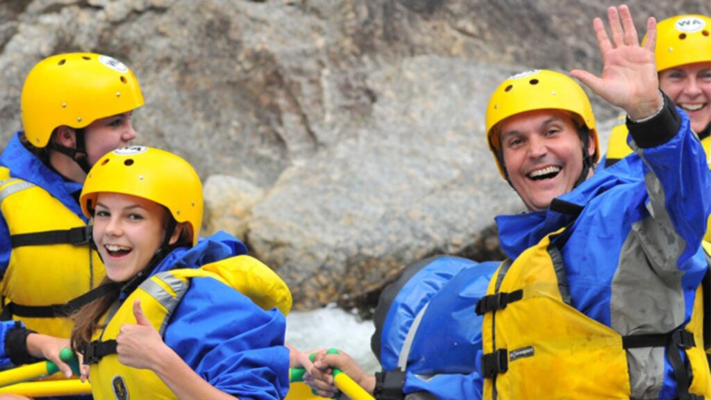 A family smiles and waves from a raft with green helmets and PFDs