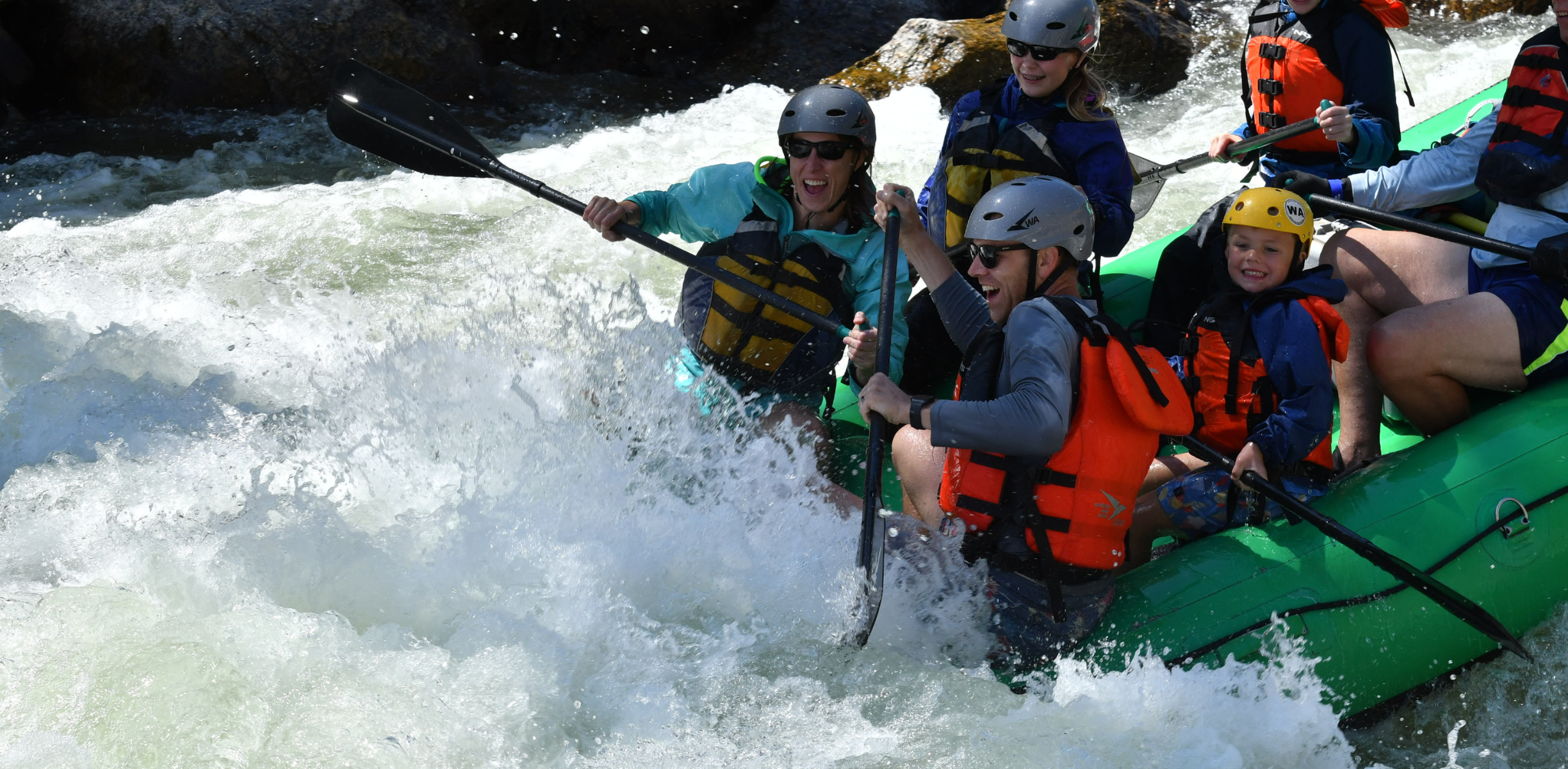 Two people on a green raft in white water hold up their paddles and smile